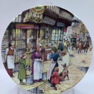 May include: A decorative plate showing a detailed street scene with shops and people. The plate depicts a vintage town with shops labeled "OTS SHOES", "SAM HALL", and "NEWSPAPERS & SWEETS". A dog sits on the cobblestone street. The plate is displayed on a clear stand.