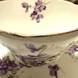 May include: White teacup and saucer set with a gold rim and purple violet floral design. The teacup has a curved handle and a scalloped edge. The saucer is also decorated with violets.