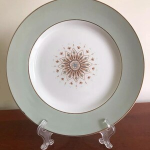 May include: A round, white plate with a pale green border and gold trim. The centre of the plate features a floral design in brown, blue, and gold. The plate is displayed on a clear plastic stand.