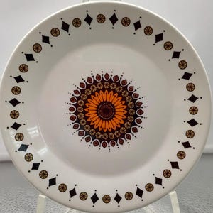 May include: White ceramic plate with a brown, orange, and red mandala design in the center. The plate has a black and gold border with a repeating diamond and circle pattern.