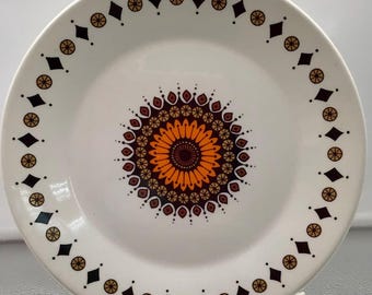 May include: White ceramic plate with a brown, orange, and red mandala design in the center. The plate has a black and gold border with a repeating diamond and circle pattern.