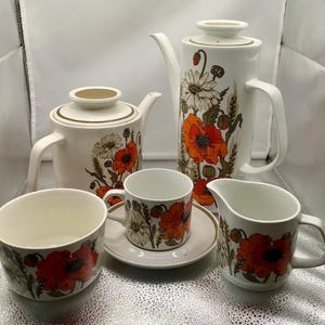Könnte beinhalten: Ein weißes Teeservice mit einem Blumenmuster mit roten Mohnblumen, grünen Blättern und weißen Blüten. Das Set beinhaltet eine Teekanne, eine Milchkännchen, eine Zuckerdose und zwei Teetassen mit Untertassen.