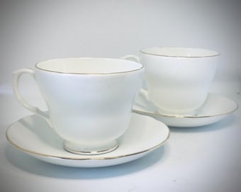 May include: Two white teacups and saucers with a gold trim. The cups have a classic shape and are perfect for enjoying a cup of tea.