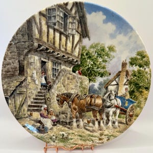 May include: Decorative plate featuring a rural scene with a horse-drawn cart, a stone cottage, and figures in period clothing. The plate has a detailed painting with a blue sky background and is displayed on a copper-colored stand.