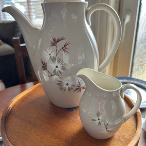May include: A grey ceramic teapot and creamer set with a floral design. The teapot has a lid and a spout. The creamer has a handle. Both pieces have a white background with brown and green floral accents.
