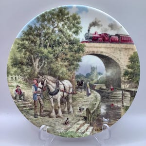 May include: Decorative plate featuring a pastoral scene with a white horse, people, and a red train on a bridge. The plate has a glossy finish and is displayed on a clear stand. The artwork depicts a rural landscape with a canal and a church in the background.