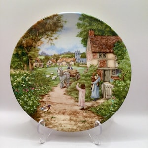 Peut inclure: Assiette décorative représentant une scène de village pastoral. L'assiette représente un chemin de terre menant à un cottage avec un toit en tuiles rouges. Une charrette tirée par des chevaux, des personnages et une église sont visibles en arrière-plan.