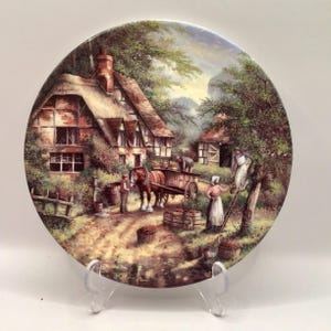 May include: Decorative plate featuring a rural scene with a thatched-roof cottage, a horse-drawn cart, and figures harvesting. The plate has a detailed, colorful painting with a pastoral theme, showcasing a vintage aesthetic.