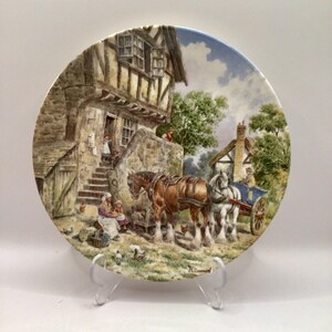 May include: Decorative plate featuring a rural scene with a horse-drawn cart, a stone cottage, and figures in traditional attire. The plate has a detailed painting with a blue sky background and is displayed on a clear stand.