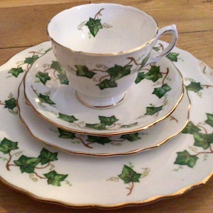 May include: A white teacup and saucer set with a gold trim and a green ivy pattern. The set includes a teacup, saucer, and a larger plate.
