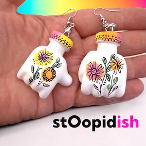 Può includere: Un paio di orecchini bianchi a forma di mano con disegni floreali. Gli orecchini presentano illustrazioni di fiori rosa, gialli e neri con foglie verdi. La parte superiore di ogni orecchino è dipinta con accenti rosa e gialli e hanno ganci argentati.