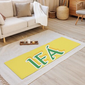 May include: A yellow yoga mat with the word "LEA" written in green letters. The mat is on a white rug in a room with a couch and a wooden floor.