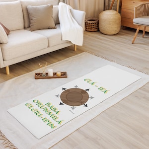 May include: White yoga mat with green and yellow text and a brown circular design with arrows. The text reads "ELA-IFA" and "BABA ORUNMILA ELERI-IPIN". The mat is on a light-colored rug.