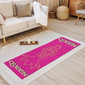 May include: A pink yoga mat with a gold outline of a woman in a yoga pose. The words "Queen" are written in gold on the mat.