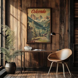 May include: A framed vintage travel poster with the word "Colorado" in red script. The poster depicts a mountain landscape with a river and trees. A wooden desk with books and a chair are in front of a wood-paneled wall.