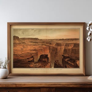 May include: A framed vintage print depicting a canyon landscape with reddish-brown rock formations under a pale sky. The print is in a wooden frame and rests on a wooden surface. The text at the bottom reads: "THE KAIBAB CANYON AT THE FOOT OF THE TOROWEAP-LOOKING EAST."