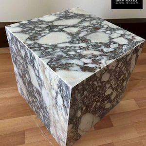 May include: A cube-shaped marble table with a unique pattern of white, gray, and burgundy veining. The table has a polished finish and is set on a wooden floor. The logo "MKM MARBLE NATURE TO LUXURY" is in the top right corner.