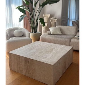 May include: A large, square coffee table with a light-colored, stone-like surface dominates the foreground. The table is surrounded by a cream-colored sofa and armchair. A tall potted plant with large green leaves adds a natural element to the room.