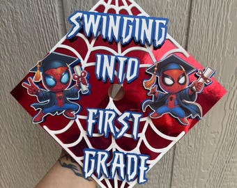 May include: A graduation cap featuring a red and white spiderweb design with the words "SWINGING INTO FIRST GRADE" in blue. Two cartoon Spider-Man figures in graduation attire are also featured.