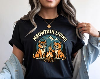 May include: Black t-shirt with a graphic featuring two orange cats camping with a campfire and mountains. The text "MEOWTAIN LIVING" arches above the image. One cat wears sunglasses and the other holds a drink.