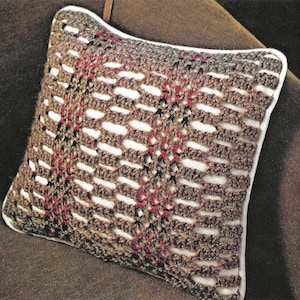 Op de afbeelding: Een vierkant gehaakt kussen met een bruin en wit open weefpatroon. Het kussen heeft een witte rand en een bruine achtergrond. Rode en zwarte accenten zijn zichtbaar in het haakpatroon. Het kussen ligt op een bruin oppervlak.