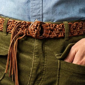 May include: A brown leather belt with a woven design and a decorative buckle. The belt has a fringe detail and is worn with green corduroy trousers and a blue shirt.