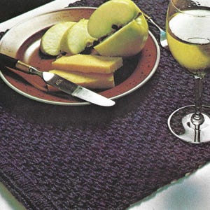 Puede incluir: Naturaleza muerta con un plato con manzanas y queso en rodajas, una copa de vino y un mantel individual de punto morado. Un cuchillo descansa sobre el plato. La copa de vino está llena de un líquido claro.