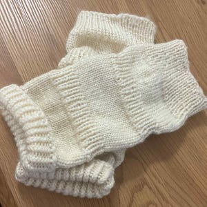 Puede incluir: Un par de calentadores de piernas de punto de color crema. Los calentadores de piernas están hechos de un hilo grueso y texturizado con un puño acanalado en la parte superior e inferior. Los calentadores de piernas están doblados uno sobre otro sobre una superficie de madera.