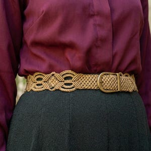 Può includere: Una cintura in macramè beige con fibbia in ottone. La cintura presenta un design intrecciato con motivi geometrici. La cintura è indossata sopra una gonna verde scuro e una camicia bordeaux.