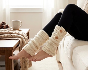 May include: Cream-colored knitted leg warmers with two wooden buttons. The leg warmers are ribbed and worn over black leggings. The image shows a person relaxing on a white couch, with a coffee table and a window in the background.