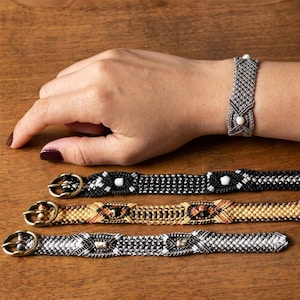 Könnte beinhalten: Makramee-Armbänder in Schwarz, Gold und Grau mit aufwendigen Webmustern und Metallverschlüssen. Ein Armband wird am Handgelenk getragen, um das Design zu zeigen. Präsentiert auf einer Holzoberfläche.