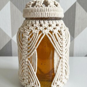 Pode incluir: Um frasco de vidro transparente com uma embalagem e tampa de macramé bege. O frasco é preenchido com um líquido dourado. O design de macramé apresenta um padrão tecido em torno do frasco e na tampa.