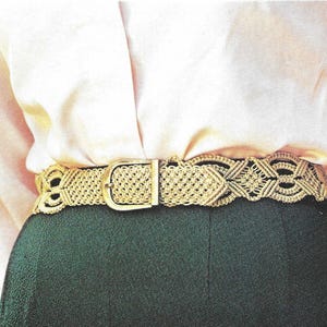 Può includere: Una cintura dorata intrecciata con una fibbia rettangolare dorata. La cintura presenta un design a trama aperta dettagliato con motivi geometrici. La cintura è indossata sopra una gonna verde scuro e una camicetta chiara.