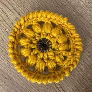 Può includere: Un sottobicchiere fatto a mano, all'uncinetto giallo, a forma di girasole. Il sottobicchiere presenta un centro marrone scuro e petali gialli dettagliati, appoggiato su una superficie di legno. Questo oggetto decorativo è perfetto per aggiungere un tocco di fascino fatto a mano.
