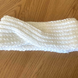 May include: A white knitted headband with a twisted design. The headband is made of a soft, textured yarn. The item is shown on a wooden surface. The headband is designed to keep the ears warm.