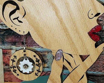 May include: Wooden earring with a floral design. The earring is round with a light wood frame and a white flower in the center. The earring is hanging from a wooden display shaped like a woman's face. The background is a brick wall.
