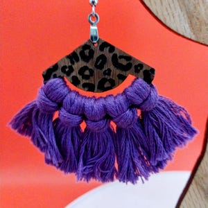 Puede incluir: Pendientes de borla morados con un colgante de madera con estampado de leopardo. Los pendientes cuelgan de ganchos plateados.