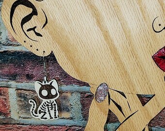 May include: A pair of Halloween-themed earrings featuring a skeleton cat design. The earrings are white with black details and hang from silver-colored hooks. The cat skeleton earrings are displayed near a wooden cutout of a face.