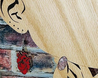 Pode incluir: Silhueta de madeira de um perfil humano com um brinco vermelho em forma de coração. O brinco é um coração anatómico detalhado. O fundo é uma parede de tijolos.