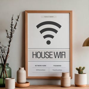 Op de afbeelding: Een ingelijste print met een wifi-symbool en de woorden "HOUSE WIFI". Hieronder staan velden voor de netwerknaam en het wachtwoord. De print zit in een licht houten lijst en de achtergrond is wit. Decoratieve items zijn een groene glazen vaas met takken en kleine keramische potten.