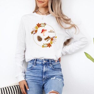 Puede incluir: Camiseta blanca de manga larga con un gráfico de un hongo, una piña, palitos de canela y hojas de otoño en una corona con el texto "hello autumn".