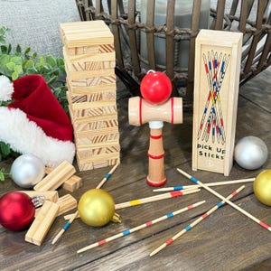 Op de afbeelding: Een verzameling houten spellen en kerstversieringen op een houten oppervlak. Inclusief een Jenga-toren, een rode kerstmuts, een rode bal, een houten kendama-speelgoed, een doosje plukstokken en diverse kerstballen.