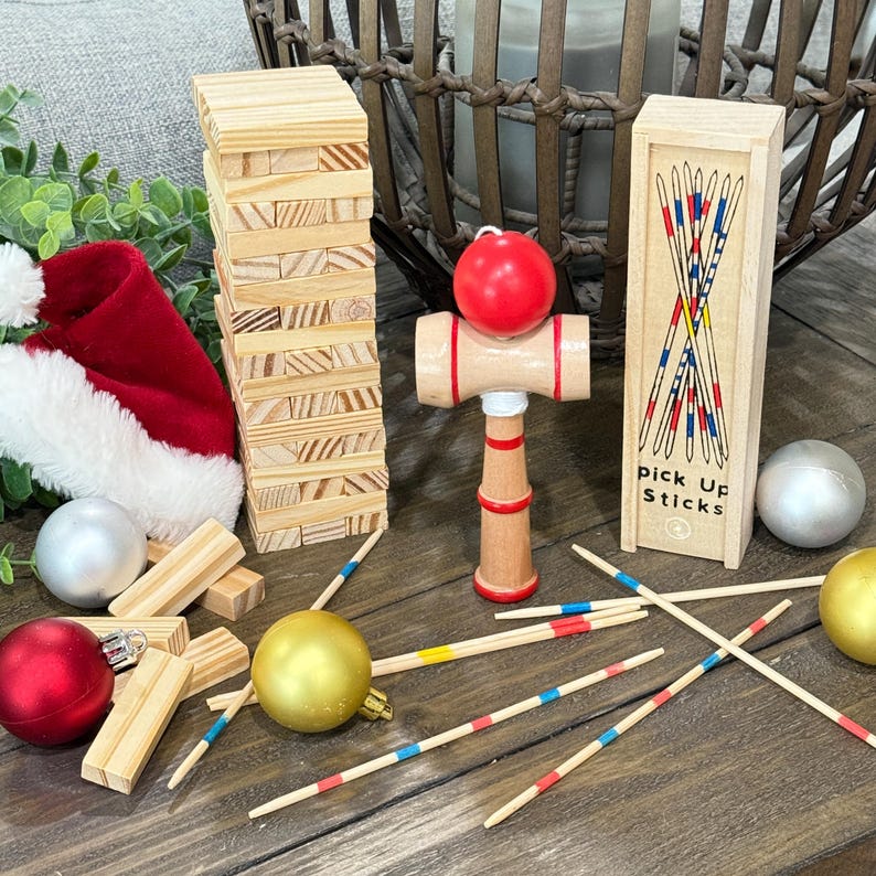 Op de afbeelding: Een verzameling houten spellen en kerstversieringen op een houten oppervlak. Inclusief een Jenga-toren, een rode kerstmuts, een rode bal, een houten kendama-speelgoed, een doosje plukstokken en diverse kerstballen.