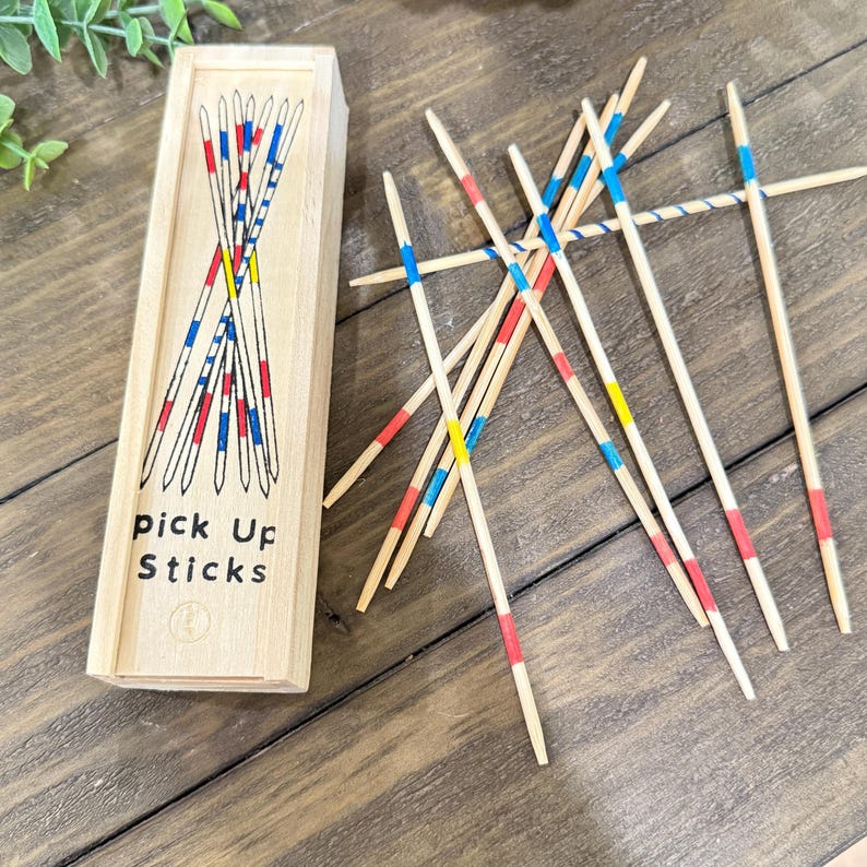Op de afbeelding: Houten mikadospel met een rechthoekige houten doos. Op de doos staat "pick up sticks". De stokken zijn beschilderd met rode, blauwe en gele banden. Gezelschapsspel.