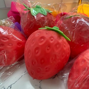 May include: Several large, bright red strawberry-shaped objects with green plastic stems are visible, each individually wrapped in clear plastic. The strawberries have a textured surface with small indentations, and are arranged on a white marble-like surface.
