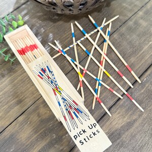 Op de afbeelding: Houten mikadospel met een houten doos. De stokken zijn beschilderd met rode, blauwe en gele banden. Op de doos staat "Pick Up Sticks". Het spel ligt op een houten oppervlak.