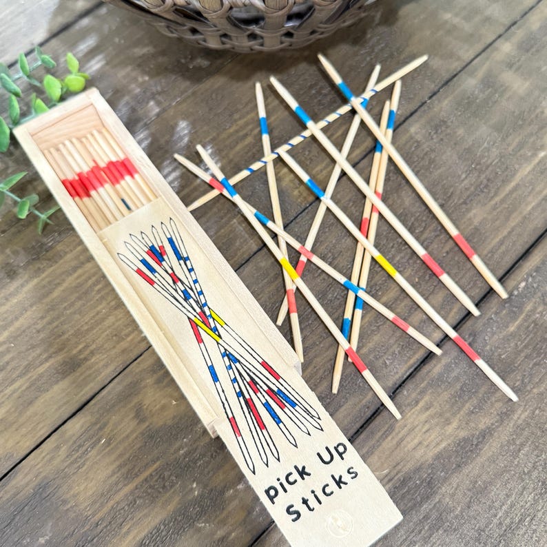 Op de afbeelding: Houten mikadospel met een houten doos. De stokken zijn beschilderd met rode, blauwe en gele banden. Op de doos staat "Pick Up Sticks". Het spel ligt op een houten oppervlak.