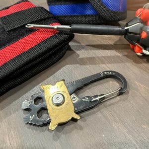 May include: A multi-tool with a gold-colored owl-shaped piece, a black carabiner, and a screwdriver. The multi-tool has various measurement markings in inches and other functions. A black and red pouch is in the background.