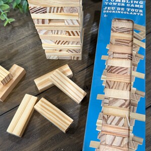 Op de afbeelding: Een houten tumbling tower game met een blauwe doos. Het spel bevat rechthoekige houten blokken, sommige gestapeld in een toren, en andere verspreid in de buurt. De doos leest "Tumbling Tower Game" en "Jeu de Tour Degringolante".