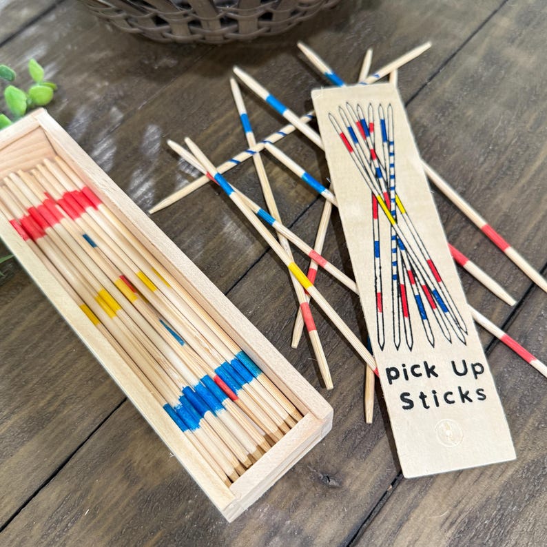 Op de afbeelding: Een houten mikadospel. De set bevat een houten doos gevuld met stokken, elk beschilderd met rode, blauwe en gele banden. De spelinstructies staan op een kaart. De stokken liggen verspreid op een houten oppervlak.